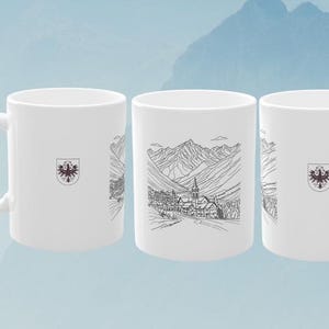 Könnte beinhalten: Drei weiße Keramikbecher mit schwarzen Strichzeichnungen eines Bergdorfes und eines Wappens. Die Becher werden vor einem hellblauen Hintergrund mit einer Bergkette präsentiert.