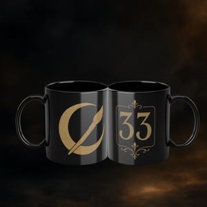 May include: Two black ceramic mugs. One mug features a gold crescent moon design with a paintbrush. The other mug has the number 33 in gold within a decorative frame. Both mugs have a black handle.