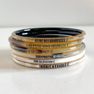 May include: A stack of six horn bangles with black lettering. The bangles feature the phrases "Reine des Boudeuses", "Raleuse mais heureuse", "Pipelette de compet'", "Bodypositive", "Sun Salutation", and "Inhale & Exhale".