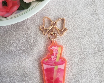 May include: A clear acrylic keychain featuring a pink and red bubble tea design with a cow on top. The keychain has a gold bow-shaped clasp and a gold ring. The background is a white, textured surface.