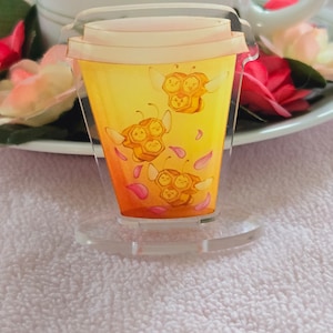 May include: A clear acrylic keychain featuring a yellow and orange cartoon coffee cup with three cartoon bees with honeycombs on their bodies. The bees are flying around the cup and there are pink flower petals around them.