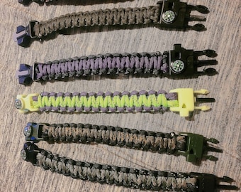 May include: Six different paracord bracelets with a compass and a clasp. The bracelets are in various colors, including red, white, blue, green, brown, and black.