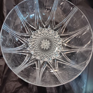 Op de afbeelding: Een heldere glazen serveerschaal met een sterrenpatroon. De schaal heeft een centraal stervormig patroon omgeven door stralende lijnen en een getextureerde basis. De ronde schaal is gemaakt van kristal of hoogwaardig glas.