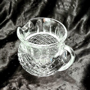 May include: A clear glass creamer with a diamond-cut pattern. The creamer has a spout for easy pouring and a curved handle. The design features a textured, patterned exterior and a smooth interior.