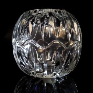 May include: A clear, round crystal bowl with a textured, cut-glass design. The bowl has a wavy rim and sits on a reflective surface, creating a mirrored effect. The bowl is a decorative item.