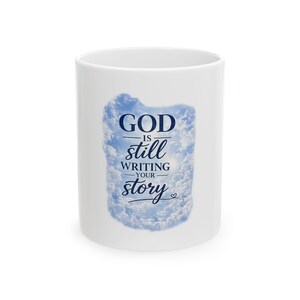 May include: A white ceramic mug featuring a central graphic of a blue sky filled with white clouds. The text "GOD IS still WRITING YOUR story" is printed in dark blue lettering over the cloud image, accompanied by a small heart symbol.