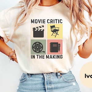 May include: Ivory-colored t-shirt with the text "MOVIE CRITIC IN THE MAKING" and film-related graphics. The design includes a film clapboard, director's chair, film reel, and notepad with a pen. The shirt is worn by a person.