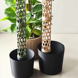 May include: Two cylindrical plant support poles with a hollow honeycomb lattice structure, one in olive green and one in beige, each standing upright inside a plain black cylindrical plant pot. Several small olive green plastic clips lie nearby.