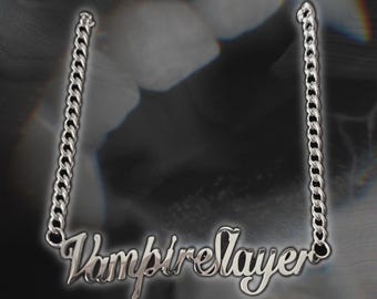 May include: A silver-colored necklace with the word "Vampire Slayer" in a cursive font. The necklace hangs from a chain with a curb link design. The background is a blurred, dark image with a hint of a face.