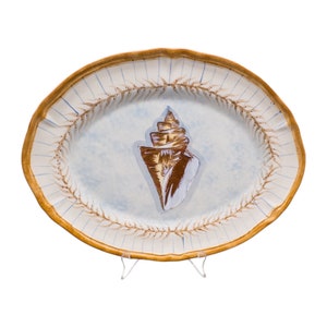 Könnte beinhalten: Eine weiße ovale Keramik-Servierplatte mit einem braunen Muschel-Design. Die Platte hat einen hellblauen Hintergrund und einen braunen Rand mit einem dekorativen Muster.