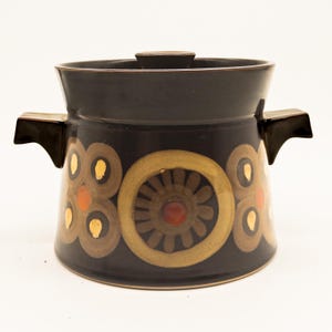 Puede incluir: Una olla de cerámica de color marrón oscuro con tapa a juego y dos asas laterales. El exterior presenta un patrón repetitivo de grandes motivos florales circulares en tonos beige y marrón con acentos naranjas y amarillos en la base.