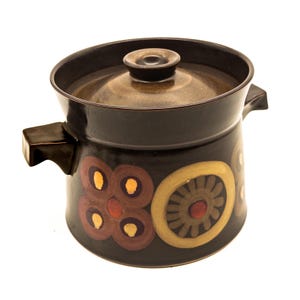 Op de afbeelding: Een donkerbruine keramische braadpan met bijpassend deksel en twee handgrepen aan de zijkanten. De buitenkant is voorzien van een decoratief patroon met cirkelvormige en bloemmotieven in tinten beige, oranje en bruin op een donkerbruine achtergrond.