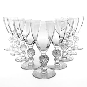 May include: A collection of clear glass goblets arranged in a semi-circle. Each goblet features a decorative, textured ball on the stem and a wide, circular base. The glasses are empty and reflect the light, creating a sparkling effect.