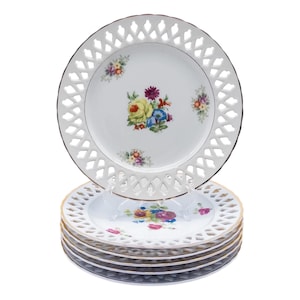 May include: A stack of decorative porcelain plates with a white openwork border and a gold rim. Each plate features a floral design with yellow, blue, and purple flowers. The plates are stacked on a white surface.