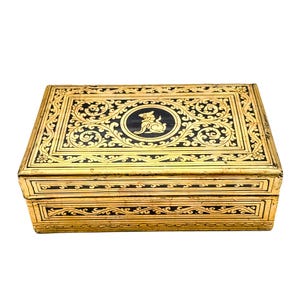 Puede incluir: Caja rectangular dorada con detalles decorativos negros. La caja presenta intrincados patrones florales y un diseño ovalado central con fondo negro y una figura de animal blanco. Probablemente utilizada para almacenamiento.