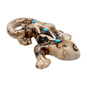 May include: A tan-colored lizard figurine with black veining and turquoise accents. The reptile sculpture has a detailed design, with a curved tail and small legs. The lizard's body is decorated with three turquoise stones.