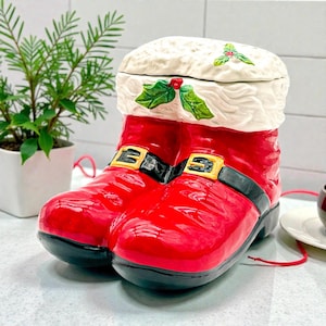 May include: A ceramic Christmas cookie jar shaped like Santa Claus's boots. The boots are glossy red with black soles and straps with gold buckles. The top of the jar is white with holly and berry accents.