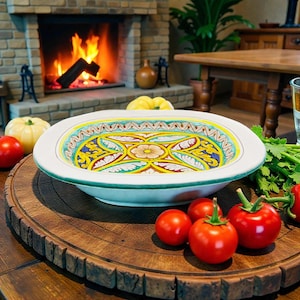 May include: A white, oval ceramic serving dish with a colorful floral design. The dish has a green rim and is placed on a wooden serving board with fresh red tomatoes. The background includes a fireplace with a burning fire.
