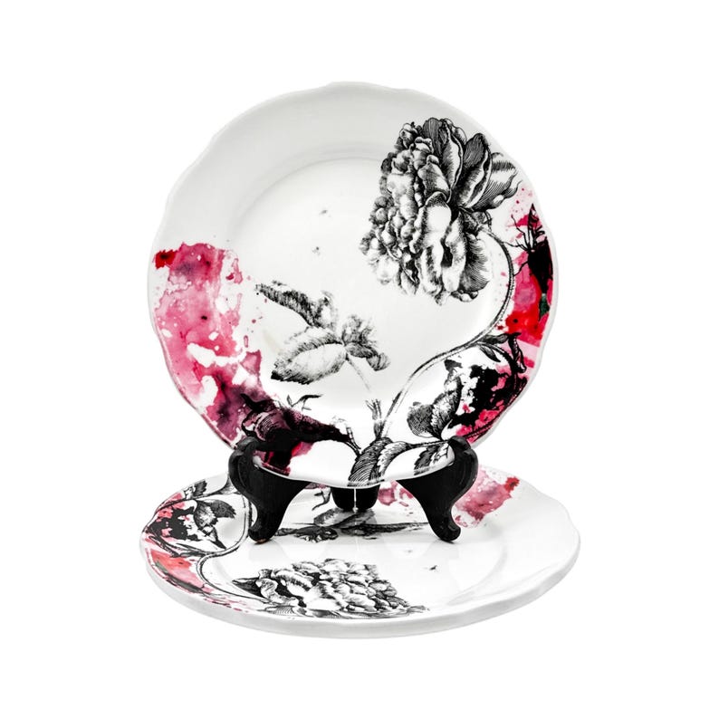 K&ouml;nnte beinhalten: Zwei wei&szlig;e Keramikteller mit floralem Design. Die Teller zeigen eine schwarz-wei&szlig;e Pfingstrosen-Illustration mit roten und rosa Aquarellakzenten. Ein Teller wird auf einem schwarzen St&auml;nder pr&auml;sentiert.