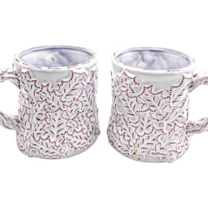 May include: Two white ceramic mugs with a textured floral design and twig-like handles. The mugs have a rustic, handmade appearance, with a red dotted pattern and a touch of gold accents. Ideal for coffee or tea.