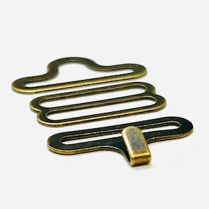 May include: A gold-toned metal bra strap hook and eye closure. The hook is a simple, rectangular shape with a rounded end. The eye is a more complex shape with multiple loops.