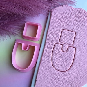 May include: Pink plastic cookie cutters in the shapes of a square, a U-shaped arch, and a combination of the two. The cutters are shown on a pink surface with the shapes cut out.