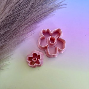 May include: Two pink plastic flower-shaped cookie cutters. The larger cutter has five petals and the smaller cutter has four petals.