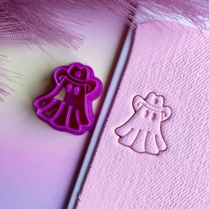 May include: Purple plastic cookie cutter shaped like a ghost wearing a cowboy hat. The cutter is shown next to a pink impression of the cutter in a pink clay-like substance.