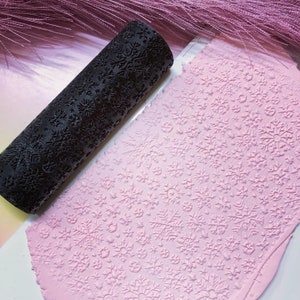 May include: A black roller with a snowflake pattern is rolling over a pink surface with a snowflake pattern. The roller is used to create an embossed snowflake design on the pink surface.