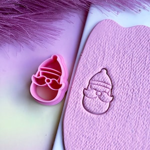May include: Pink plastic cookie cutter shaped like a cartoon Santa Claus with a hat and beard. The cutter is shown next to a pink impression of the cutter in a sheet of pink modelling clay.
