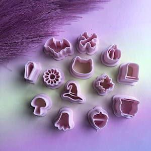 May include: A set of 12 pink plastic cookie cutters shaped like Halloween themed objects, including a bat, a pumpkin, a witch's hat, a ghost, a coffin, a cat, a skull and crossbones, a mushroom, a cauldron, an acorn, a leaf, and a flower.