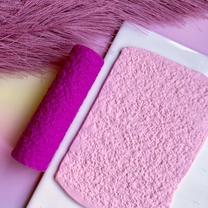 May include: A close-up of a pink textured roller and a pink textured sheet of material. The roller is made of a soft, flexible material and has a textured surface. The sheet of material is also textured and has a smooth surface.