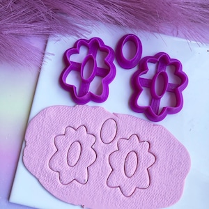May include: Purple plastic cookie cutters in the shape of flowers with a centre hole. The cutters are on a white surface with pink fondant that has been cut out with the cutters.