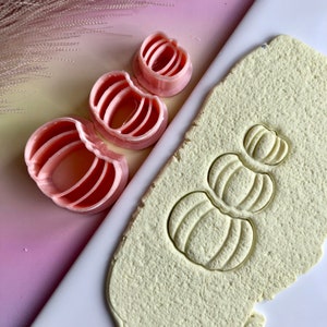 May include: Three pink plastic pumpkin cookie cutters of different sizes, with a single stack of three pumpkin shapes pressed into a light yellow dough.