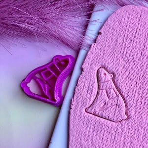 May include: A purple plastic cookie cutter shaped like a polar bear. The cutter is used to create a polar bear shape in pink play dough.