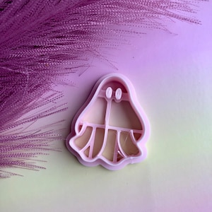 May include: Pink plastic cookie cutter shaped like a ghost with two eyes and a wide smile. The ghost has a flowing gown with a scalloped hem.