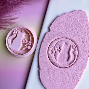 May include: A pink plastic cookie cutter shaped like a cat sitting on a crescent moon with stars. The cutter is used to create a similar design in pink play dough.