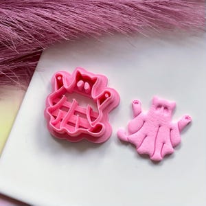 May include: Pink plastic cookie cutters shaped like a ghost and a cat wearing a ghost costume.