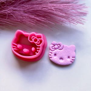 May include: Two pink Hello Kitty cookie cutters and a cut-out cookie. The cutter is a bright pink, and the cookie is a lighter shade of pink. The cutter has a bow and facial features.