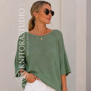 Pode incluir: Um suéter de malha verde-sálvia com um decote canoa largo, mangas soltas de três quartos e uma textura sutilmente canelada. A peça é combinada com calças brancas e inclui o texto vertical KNITORASTUDIO KNITTING PATTERNS.