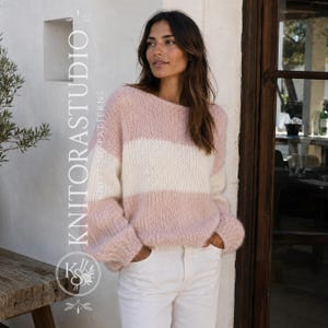 Peut inclure: Un pull en tricot duveteux à manches longues avec de larges rayures horizontales rose pâle et crème. Le pull présente une coupe décontractée avec une encolure ronde et est associé à un pantalon blanc. Le texte KNITORASTUDIO KNITTING PATTERNS est visible.