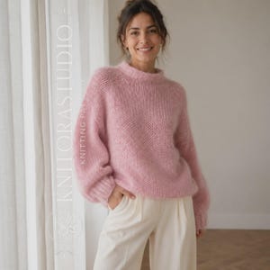 Puede incluir: Un suéter de manga larga de color rosa claro, suave y difuso, con cuello redondo y puños acanalados. El suéter presenta un patrón de tejido suelto y texturizado. El texto KNITORASTUDIO KNITTING PATTERN es visible verticalmente a la izquierda.