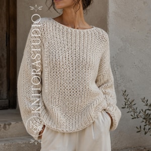 Op de afbeelding: Een gebroken witte, grofgebreide trui met een brede boothals en lange, losse mouwen. Het kledingstuk heeft overal een verticale ribbelstructuur. De tekst KNITORASTUDIO KNITTING PATTERNS staat verticaal gedrukt aan de linkerkant van de afbeelding.
