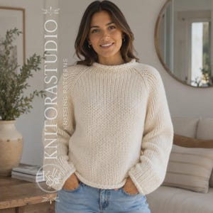 Könnte beinhalten: Ein cremefarbener, langärmeliger, grob gestrickter Pullover mit Rundhalsausschnitt und Raglanärmeln. Der Pullover hat eine vertikale Rippenstruktur und wird zu einer blauen Jeans getragen. Der Text KNITORASTUDIO KNITTING PATTERNS ist sichtbar.