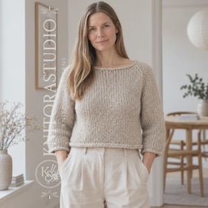 Op de afbeelding: Een beige trui met lange mouwen, een ronde halslijn en een grove breistructuur. De trui heeft een kortere lengte en wordt gecombineerd met een lichtgekleurde broek. De tekst KNITORASTUDIO is zichtbaar aan de linkerkant van de afbeelding.