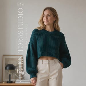 May include: A dark teal, long-sleeved, ribbed knit sweater with a cropped hemline and a crew neckline. The sweater features voluminous sleeves and is paired with light beige trousers. The text KNITORASTUDIO KNITTING PATTERNS is visible.