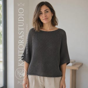 Può includere: Un maglione lavorato a maglia grigio antracite scuro con un ampio scollo a barchetta e maniche a tre quarti. Il capo presenta un motivo a maglia testurizzato e un semplice orlo arrotolato. Il testo KNITORASTUDIO KNITTING PATTERNS è visibile a sinistra.