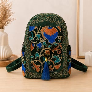 May include: A dark green velvet backpack featuring intricate gold-tone embroidered scrollwork patterns. The front pocket flap displays a vibrant floral motif in orange and blue, accented by a long, deep blue tassel hanging from the centre.