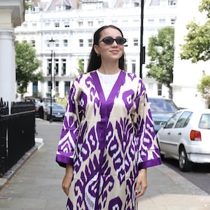 May include: A long-sleeved, open-front duster coat featuring a bold purple and cream ikat pattern. The garment has a V-neckline, wide sleeves, and a straight hemline that falls below the knees. It is worn over a plain white t-shirt and blue jeans.