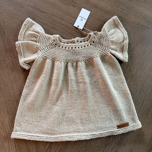May include: A light tan knitted baby dress with ruffled sleeves. The dress features a round neckline with a decorative pattern and a small wooden tag. The dress is made of a soft, textured yarn.